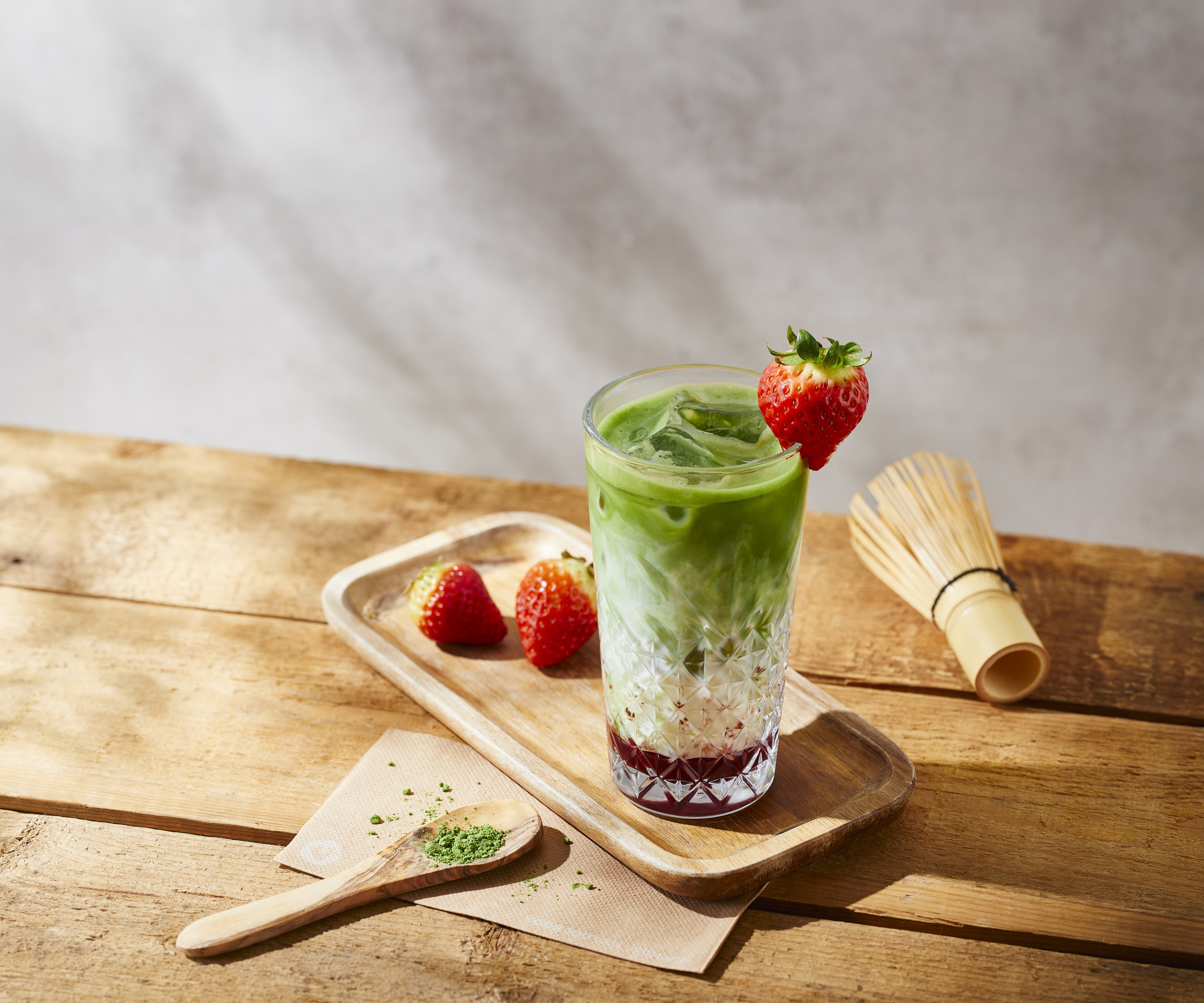 Verhage Iced Latte Matcha Strawberry met aardbeiensaus, kokosmelk, matcha en verse aardbeien op een zonnige houten tafel.